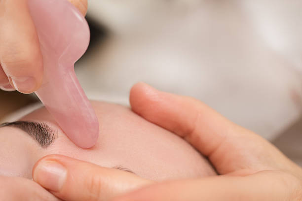 neat mujer cosmetóloga manos masajista haciendo masaje con raspador gouache de jade rosa en forma de corazón. concepto de cuidado de la piel del cuerpo de la cara, tratamiento facial antienvejecimiento. herramientas y equipos de belleza spa - spooning fotografías e imágenes de stock