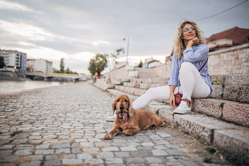 One beautiful young woman with curly blonde hair, dressed in striped shirt and white pants, sitting on the stairs near the river outdoors in the city. Her cocker spaniel dog keeping her company.