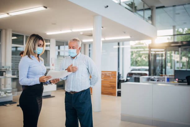 Beautiful saleswoman talking with senior man at the car dealership store Two people, young beautiful saleswoman talking with senior man at the car dealership store, they are wearing protective masks do to coronavirus. car rental covid stock pictures, royalty-free photos & images