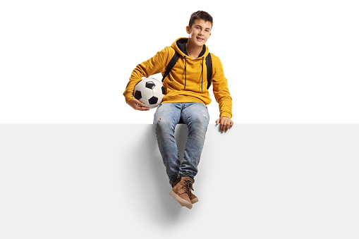 Teenager boy holding a soccer ball and sitting on a white panel board isolated on white background