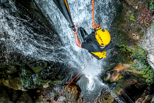 A high angle view of a male canyoning adventurist making a descent down a waterfall in a beautiful canyon and making selfie at the same time.