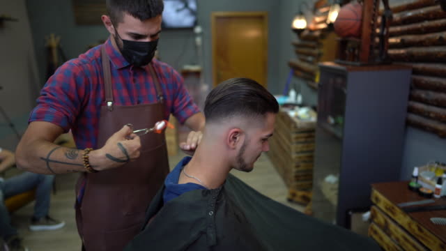 Barber with protective face mask trimming man's hair burning it by fire at barber shop