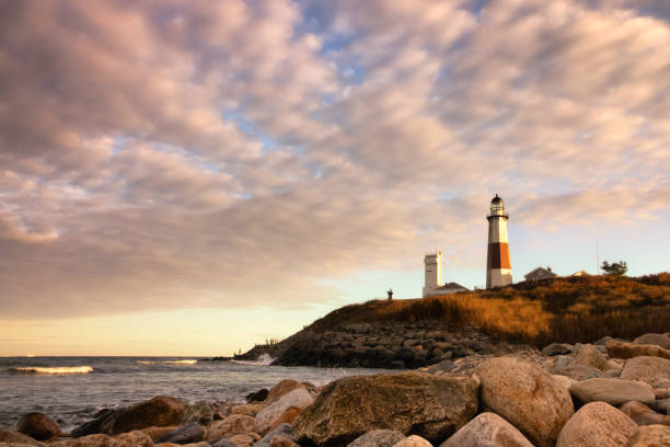 теплый золотой свет на закате, освещающий сторону маяка, сидящего на скале. монтаук-пойнт, ny - the hamptons long island lighthouse стоковые фото и изображения