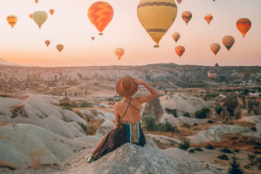 Back View Young Adult Woman Sitting On The Ground Watching Balloons Looking At The Valley. Cappadocia sunrise