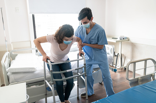 Male nurse helping female patient stand up from hospital bed while she leans on a walker both wearing protective facemasks -  Pandemic Lifestyles