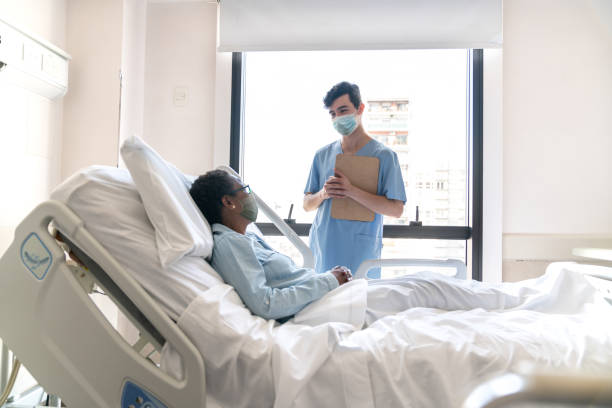 freundliche männliche krankenschwester überprüfen schwarze krankenhauspatientin auf dem bett liegend, während er eine medizinische karte hält, die beide schützende gesichtsmasken tragen - zivilist stock-fotos und bilder