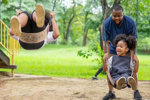 Happy African father with two little daughter playing swing together at playground in the park.