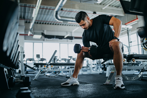 Man sitting on one gym machine and doing biceps.