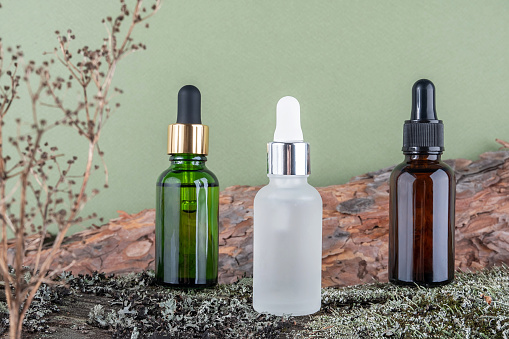 Three brown, green, white glass bottles with serum, essential oil or other cosmetic product on tree bark covered with moss against green background. Natural Organic Spa Cosmetic Beauty concept.