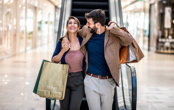 young attractive happy couple hugging, smiling and holding shopping bags while walking in shopping mall. - mercadoria imagens e fotografias de stock