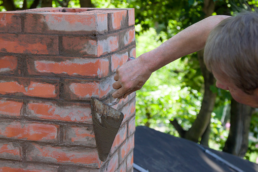A man making masonry works, working with a trowel and making a chimney of red bricks in summer