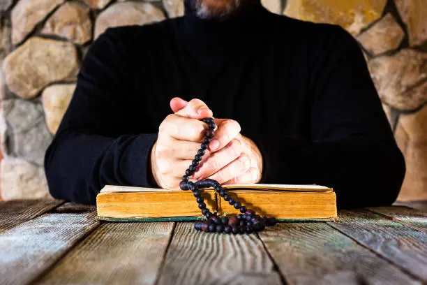 man in black clothes with a rosary in his hands in front of an open old book bible or quran, the concept of prayer