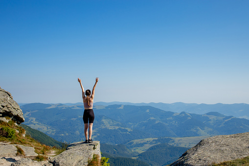 The young girl at the top of the mountain raised her hands up on blue sky background. The woman climbed to the top and enjoyed her success. Back view.