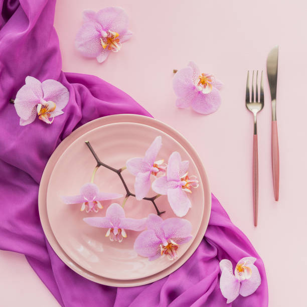 istock Flower and table settings overhead composition on light pink background. Pink ceramic plates, cutlery and perple orchid flowers. Holiday modern table decoration. Romantic date. 1277296734