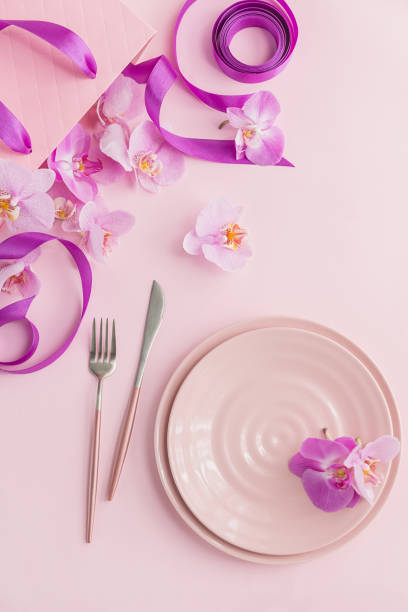istock Flower and table settings overhead composition on light pink background. Pink ceramic plates, cutlery, pink gift bag with purple ribbons and pink orchid flowers. Holiday modern table decoration 1277296740