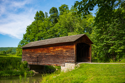 The Hyde Hall Covered Bridge, built in. 1825 and is the oldest existing covered bridge in the U.S., rests at the end of a dirt road during a summer day.