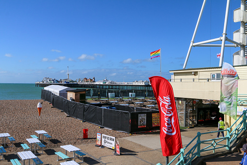 Rainbow Flag on Brighton Beach in East Sussex, England, with a Coca-Cola banner also visible in front of the Palace Pier. People can be seen in the background.