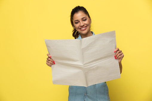 Positive casual woman smiling and reading newspaper while standing on yellow studio background
