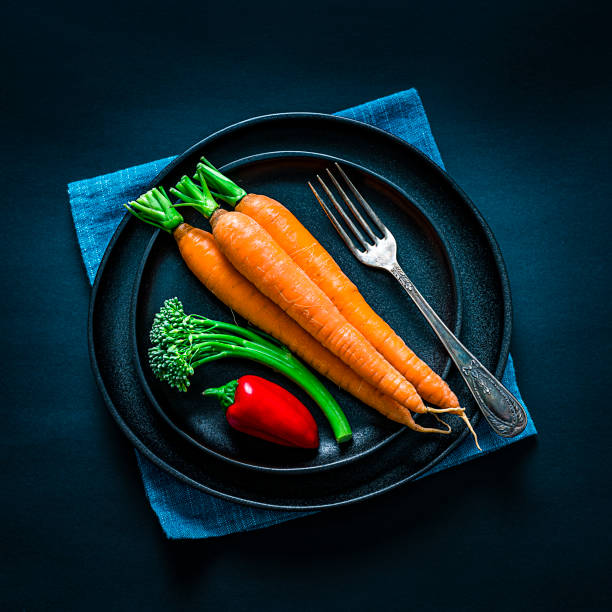 Vegan diet concept: overhead view of a black plate with fresh organic raw carrots, broccoli and red pepper. The plate is on a blue textile napkin. Predominant colors are orange, green, red and black. High resolution 28Mp studio digital capture taken with SONY A7rII and Zeiss Batis 40mm F2.0 CF lens