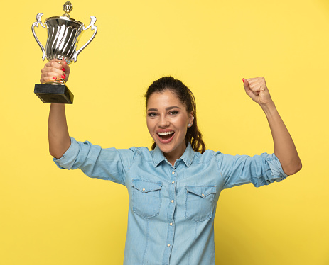 Cheerful casual woman holding trophy and celebrating, shouting while standing on yellow studio background