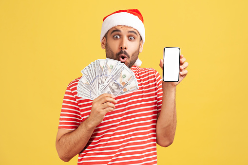 Surprized wondered man in santa claus hat holding empty blank smartphone and fan of dollars in hands, looking at camera with big eyes and open mouth. Indoor studio shot isolated on yellow background