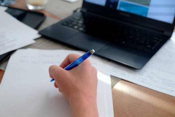 Cropped shot of young Caucasian male hands writing notes on a paper, in front of his laptop during homeschooling amidst Covid 19 pandemic