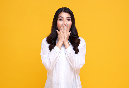 Surprised and Shocked asian woman covering mouth with hands isolated on bright yellow background.