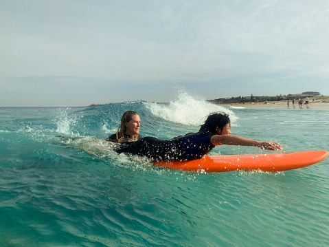 A Middle Aged Chinese woman is wearing a wetsuit and surfing through the waves on her surfboard in the sea in Australia with an instructor by her side, helping her learn.