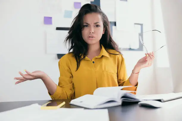 Photo of Perturbed office worker. Stressed young woman throw up her hands. Lady in yellow blouse holding glasses in hand. There is an open notebook and phone on the table. Office work / work from home concept.