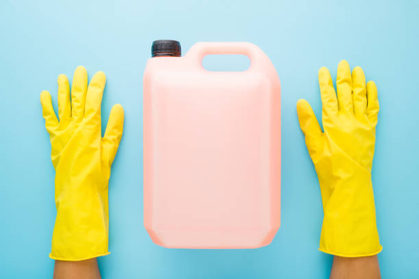 Hands in rubber protective gloves and 5 L plastic container on blue table background. Detergent for different surfaces in kitchen, bathroom and other rooms or washing liquid for dishes. Top down view.
