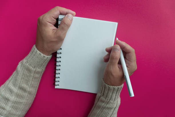 mans right hand in winter sweater hold white pen and writes his plans and list of motivation for future for new year in white papered blank spiral notebook, pink backfround - backfround imagens e fotografias de stock