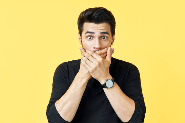 Shocked man covering mouth with hands over yellow background. Shocked man covering mouth with hands over yellow background. emotions and secret concept Hidden Meaning stock pictures, royalty-free photos & images