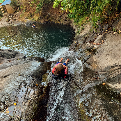 A high angle view of a young girl sliding down the Aling Aling waterfall in Bali, Indonesia into the lagoon at the bottom, where her mother is waiting for her.