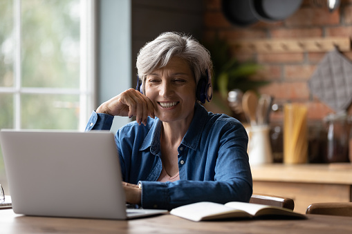 Smiling modern mature 60s Caucasian woman in earphones look at laptop screen watch webinar online at home. Happy senior grey-haired female have fun study on internet. Elderly technology concept.