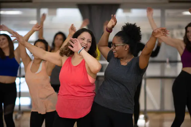 A diverse group of women smile and laugh as they dance in a fitness room.