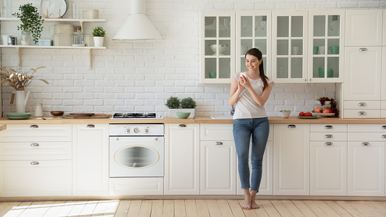 Happy woman surfing net on mobile standing barefoot on warm floor in Scandinavian kitchen. Young housewife in her 20s shopping online using smartphone leaning on modern kitchen countertop and smiling