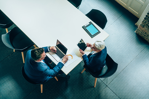 Above view of a two confident business persons sitting on a conference table in the office. Businessman and businesswoman in meeting discussing business strategy, using laptop and digital tablet. Business coworkers working together in the office.