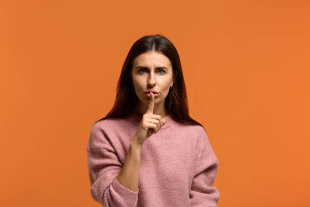 istock I demand complete silence. Serious beautiful woman in pink sweater with mysterious look, keeps index finger over lips, makes hush sign, looks at camera. on orange background 1277407048