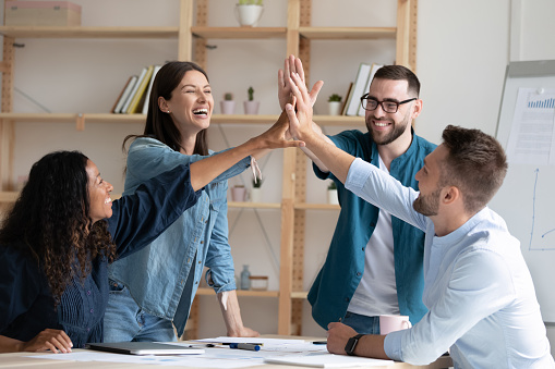 Overjoyed diverse employees giving high five at corporate meeting, excited colleagues joining hands, celebrating business achievement, good teamwork results, engaged in team building activity