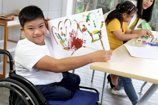 Disabled kids classroom, children having fun during study at school, kids learning together, schoolboy on wheelchair showing his painting in art classroom