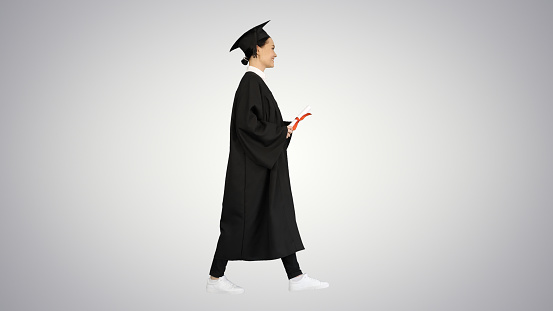 Wide shot. Side view. Happy female student in graduation robe walking and cheering with her diploma on gradient background. Professional shot in 4K resolution. 043. You can use it e.g. in your medical, commercial video, business, presentation, broadcast