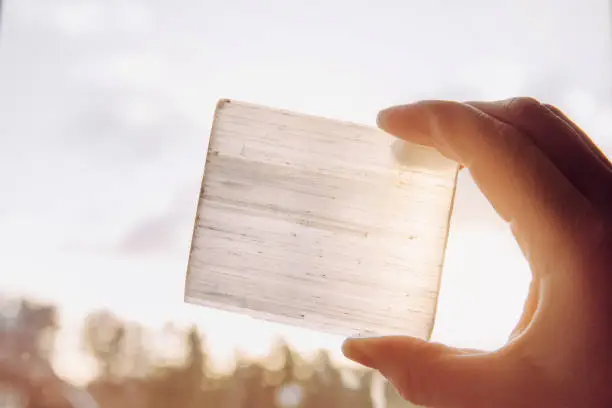 Person holding natural mineral stone Selenite plate against sun and blue sky, stone has healing and cleansing properties. Also used for recharging other mineral crystal stones.