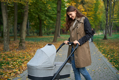 Side view of brunette woman in grey coat standing with baby stroller. Mother with baby walking in park. Watching on baby and smiling. Concept of motherhood.