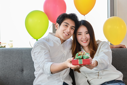 New Year, Christmas and Holiday Seasoning concept.  Portrait of asian man and woman sitting and holding gift box with colorful party balloon.