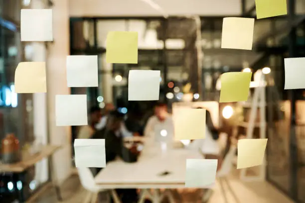 Shot of sticky notes on a glass window with businesspeople having a late night brainstorming session in a modern office