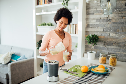 Cheerful African American woman having a healthy breakfast at home, making a shake out of different kinds of fruits and smiling