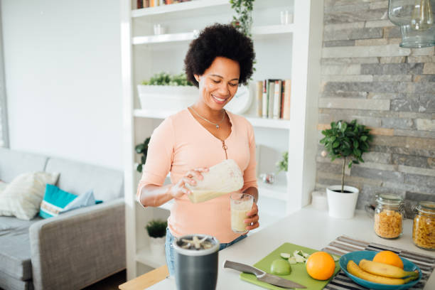Happy African American woman preparing a nutritional breakfast at home Cheerful African American woman having a healthy breakfast at home, making a shake out of different kinds of fruits and smiling protein drink stock pictures, royalty-free photos & images