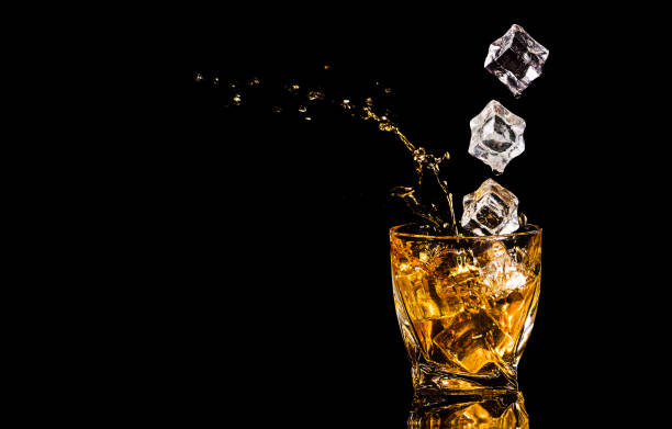 黒い背景の上に氷の立方体から飛び散ったウイスキーのグラス。アルコールが飛び散る。ウイスキーやコニャック、またはアルコールの別のタイプ - whisky alcohol pouring glass ストックフォトと画像