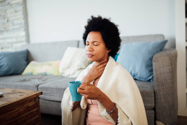 Sick African American woman staying at home African American woman, fallen ill is staying at home wrapped in a blanket socially distancing and quarantining herself, feeling her throat hurt and being sore, having a cup of hot tea sore throat stock pictures, royalty-free photos & images