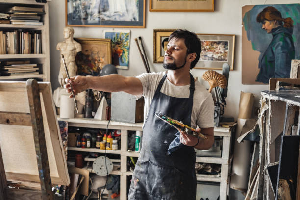 istock Male painter at art studio indoors holding paintbrush in front checking perspective looking at canvas pensive 1277421325